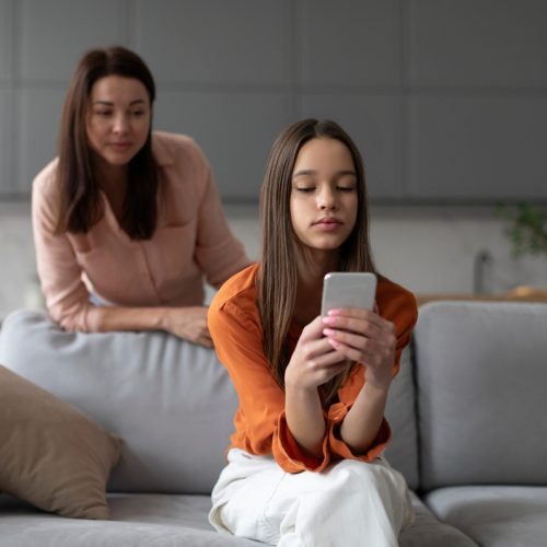Meisje zit op de bank met haar smartphone in haar handen. Haar moeder kijkt over haar schouder mee.